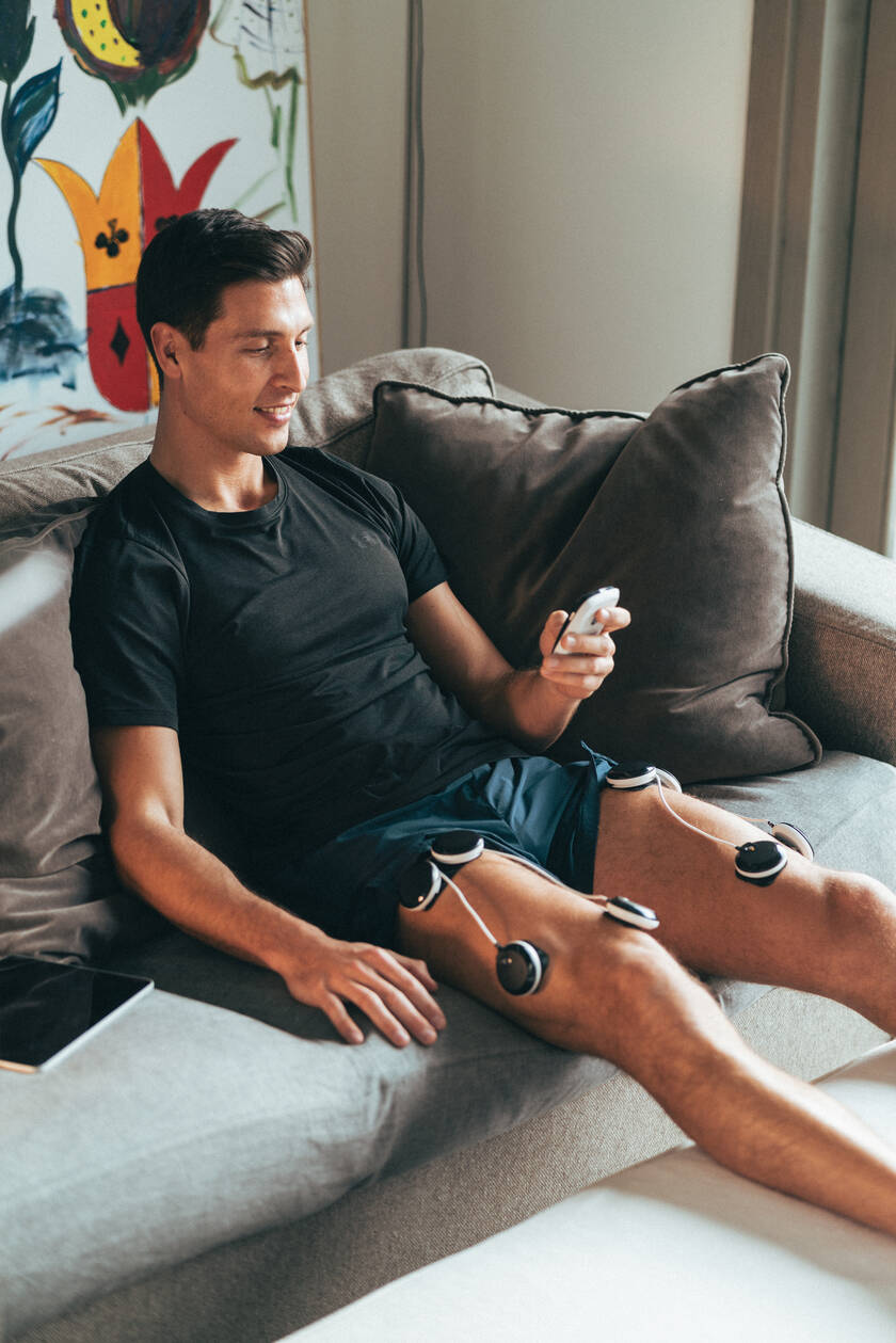 A man relaxing on a sofa, using the Compex SP 8.0 EMS muscle stimulator on his quads